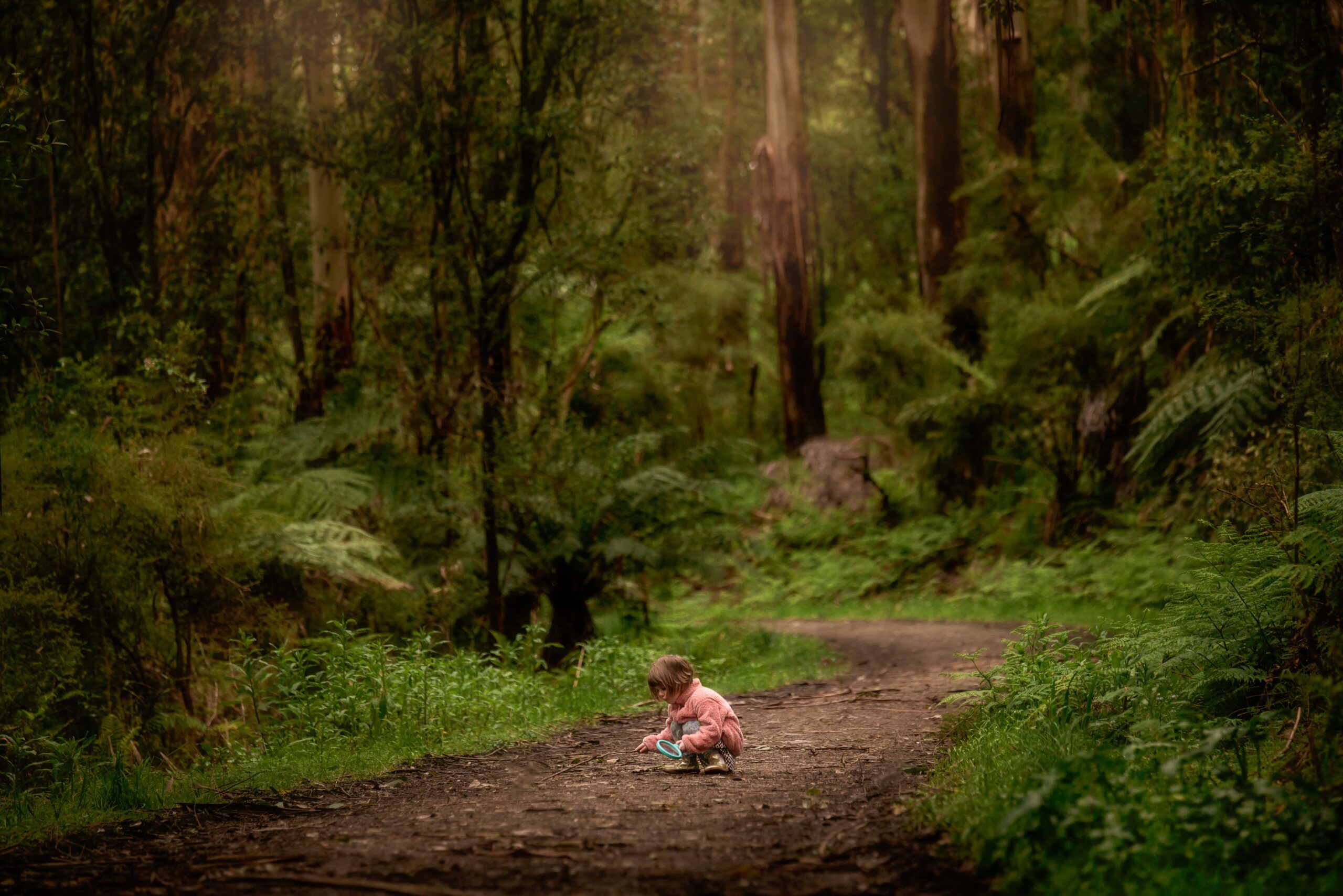 Little girl in pink jacket with a magnifying glass crouched down on a forest path