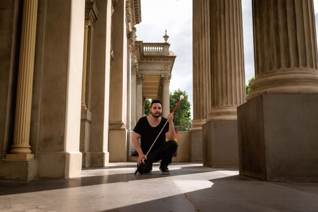 Male dressed in black with white cane crouching down amongst columns of Fitzroy Townhall with rainbow flag behind him.