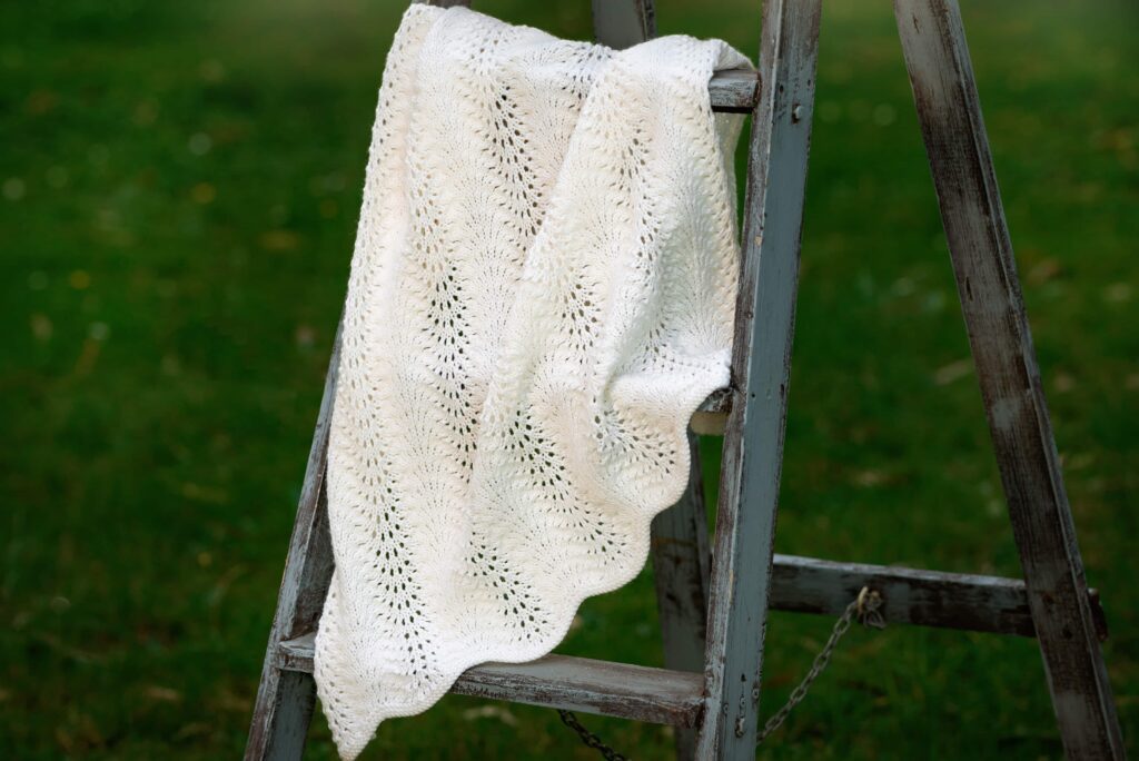 White hand knitted baby blanket with scalloped edge hanging on vintage blue timber ladder
